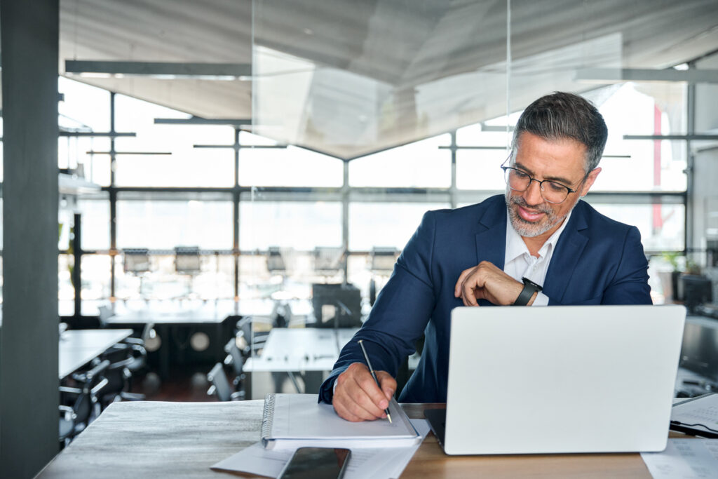 Business man working on laptop computer in office writing notes.